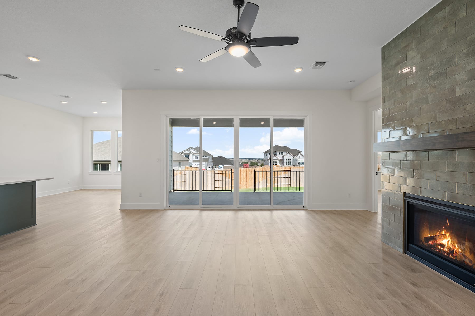 A spacious and modern living room with a fireplace, hardwood floors, and a ceiling fan, offering a view of the surrounding neighborhood through large windows.