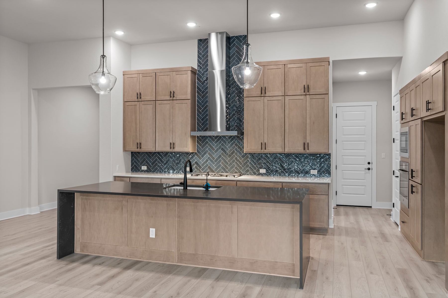 A modern and spacious kitchen with light-colored wood cabinets, a central island, and a patterned tile backsplash, illuminated by pendant lights.