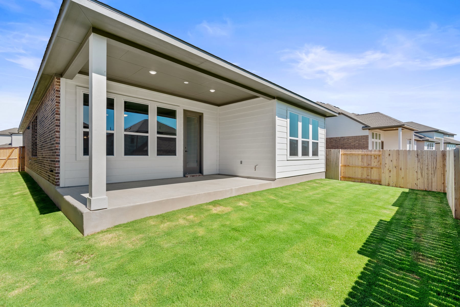 A modern, single-story house with a covered porch and large windows stands on a well-manicured lawn, surrounded by a wooden fence in the background.