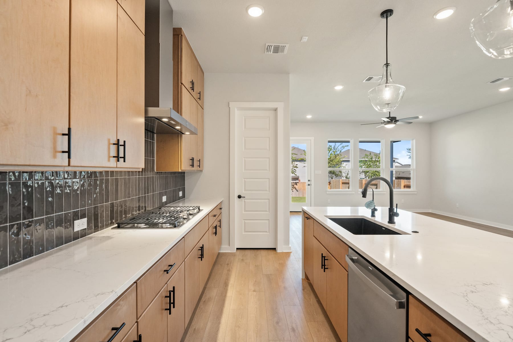 A modern, well-lit kitchen with light wood cabinets, white countertops, and a tiled backsplash, leading to a hallway with a white door.