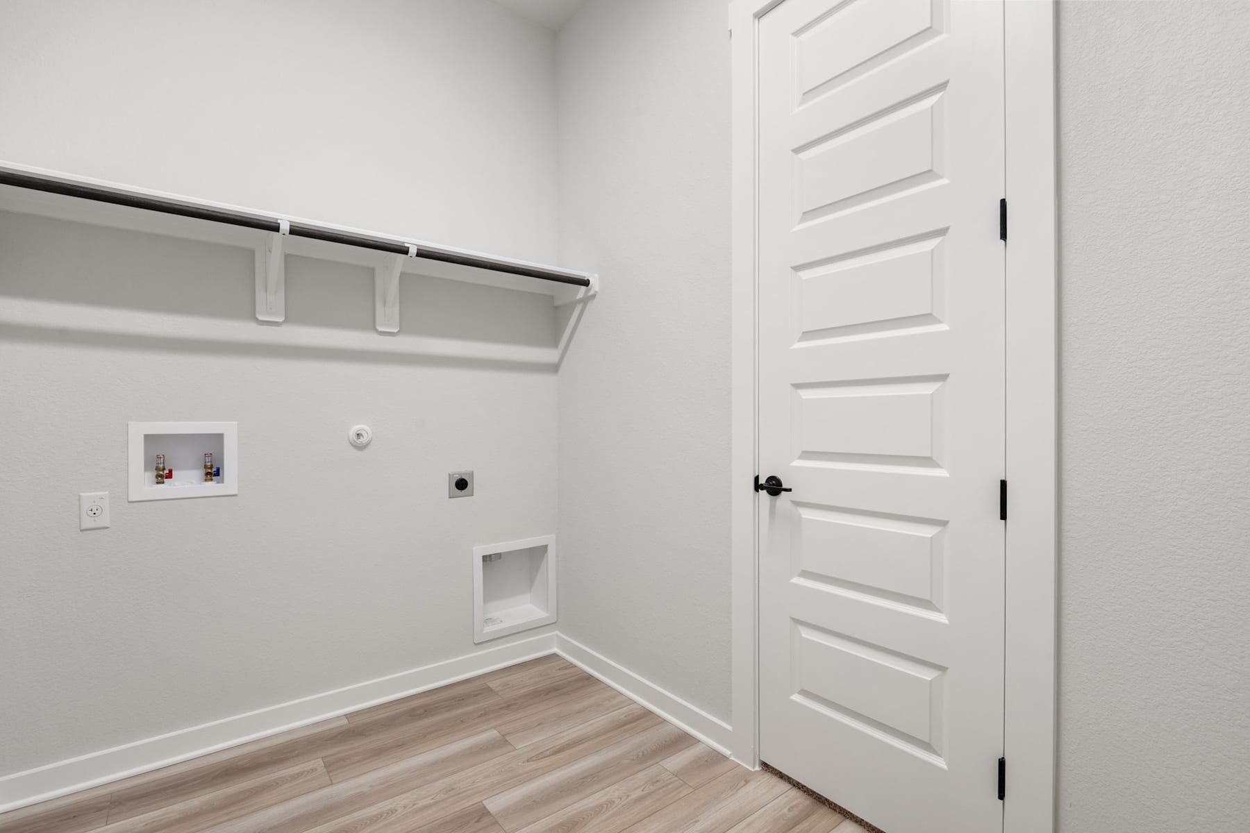 A small, minimalist room with a white door, a white wall, and a wooden floor. The room appears to have a closet or storage space with shelves visible.