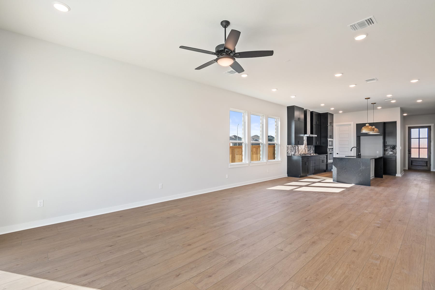 A spacious, open-concept living room with a ceiling fan, hardwood floors, and a view of a kitchen area in the background.