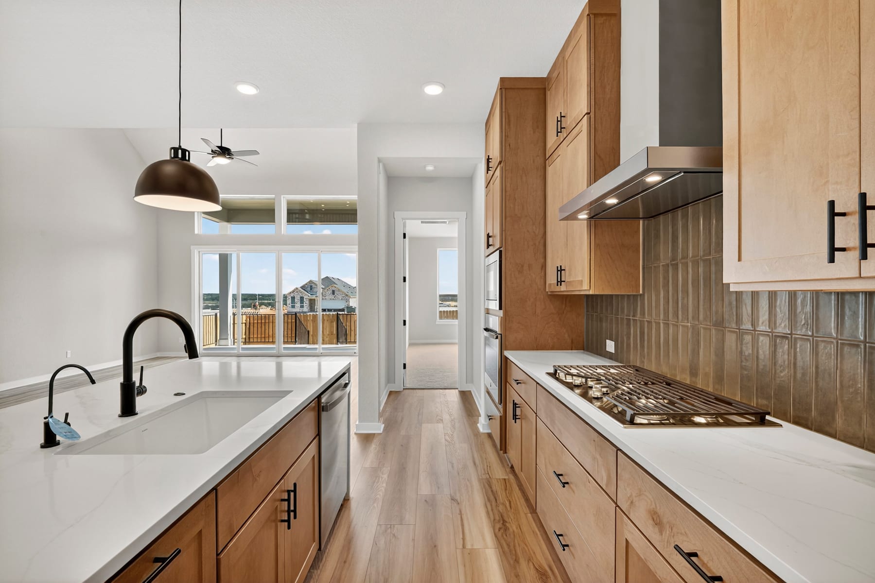 A modern, well-lit kitchen with wooden cabinets, a white countertop, and a view of the outdoors through a window at the end of the room.