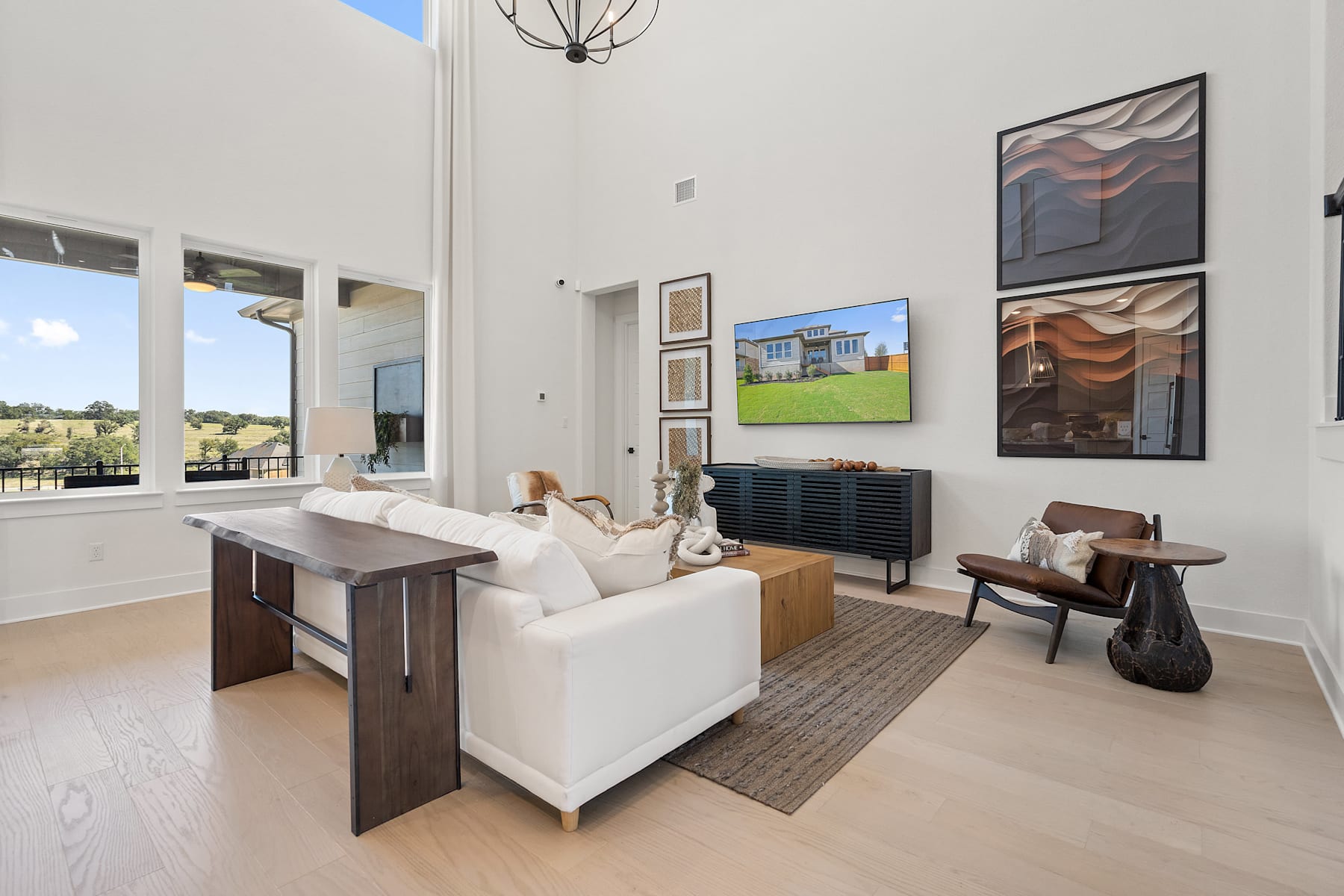 A modern and spacious living room with a white sofa, wooden furniture, and abstract artwork on the walls, set against a backdrop of large windows overlooking a scenic outdoor landscape.