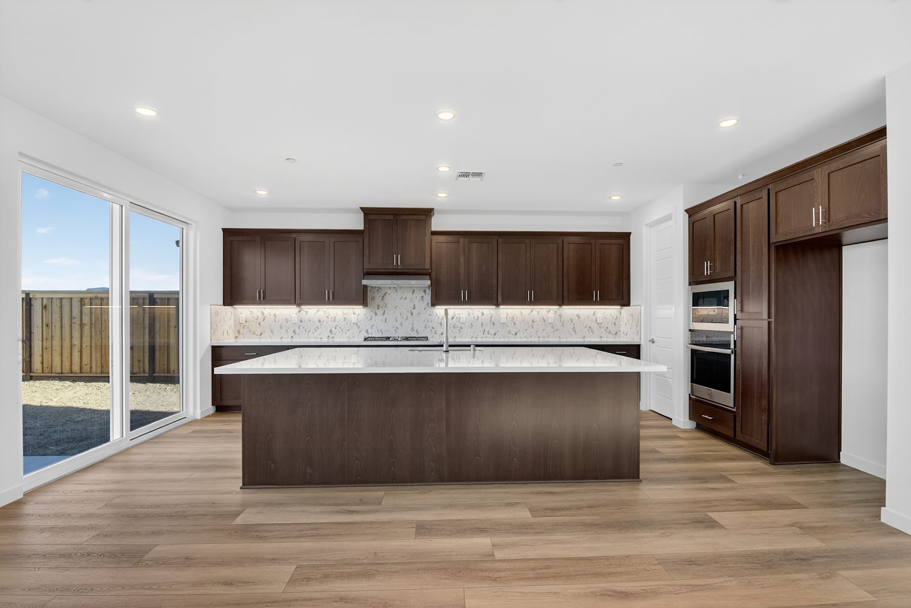 A modern, spacious kitchen with dark wood cabinets, a large island, and a view of the outdoors through a sliding glass door.