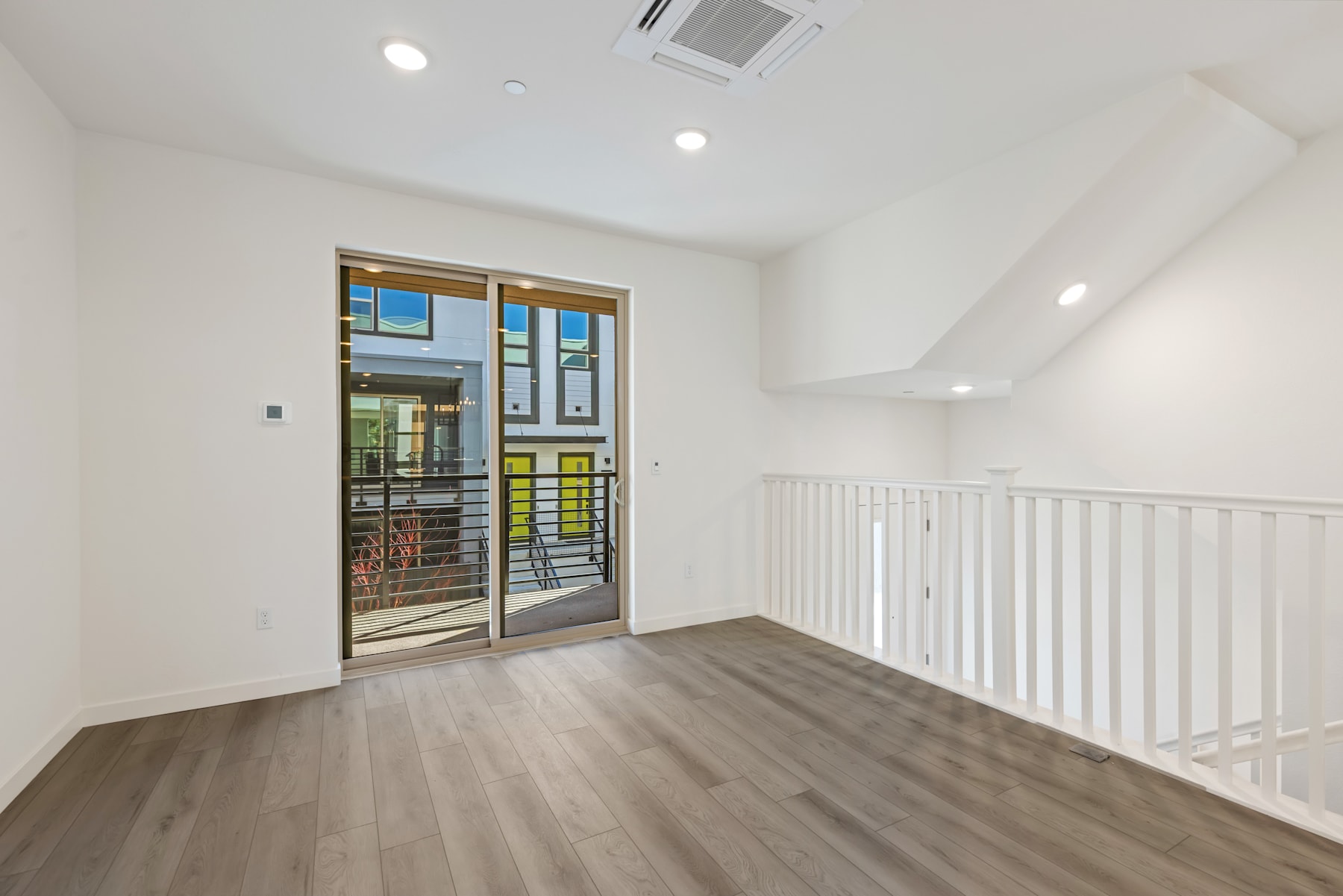 A bright and airy room with a sliding glass door leading to a balcony, hardwood floors, and a white railing along the second-floor landing.