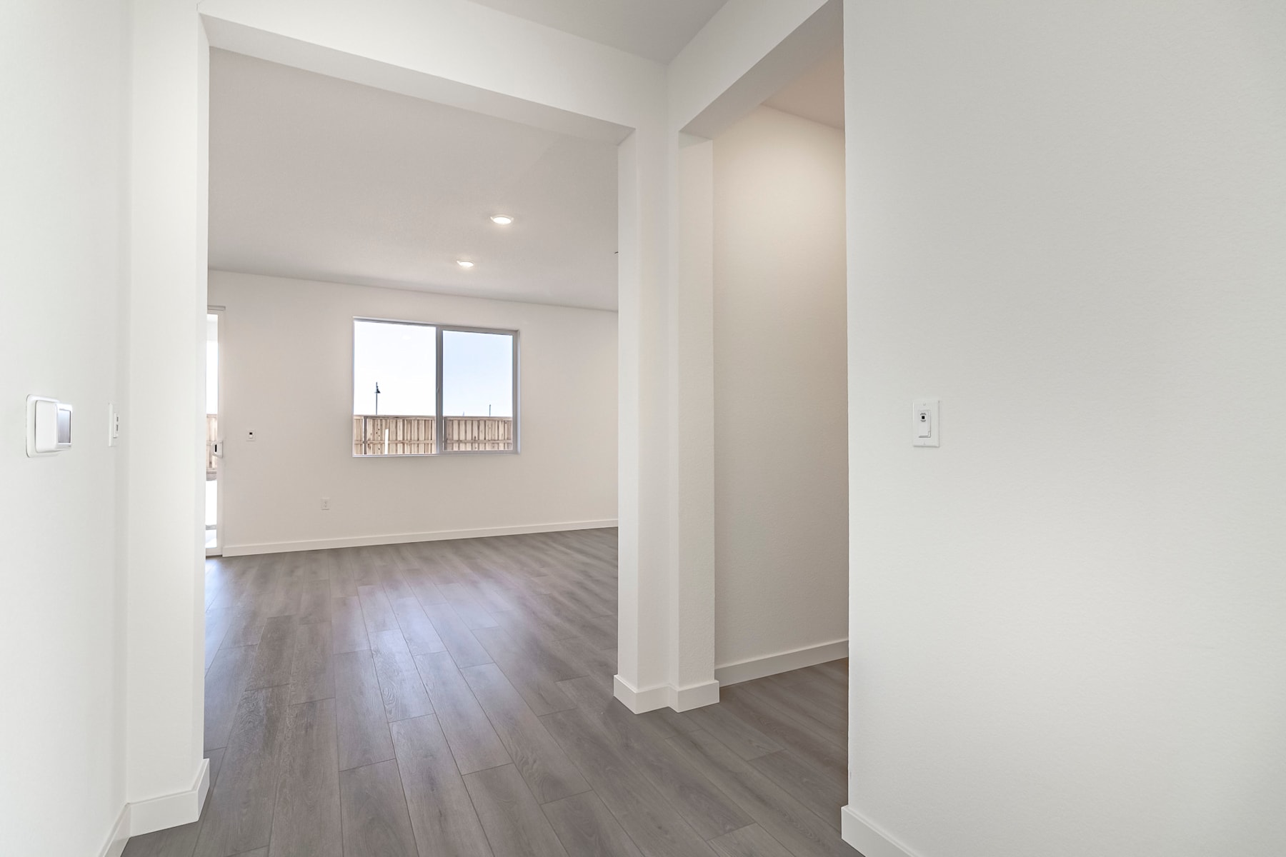 A spacious, well-lit room with white walls and a hardwood floor, leading to a window with a view of the outdoors.
