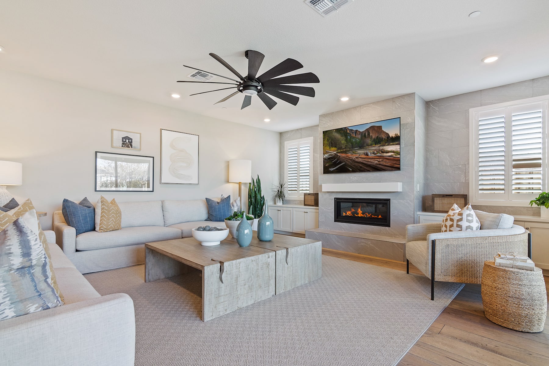 A cozy and modern living room with a large windmill-style ceiling fan, a fireplace, and various decorative elements such as artwork, plants, and textured furnishings.