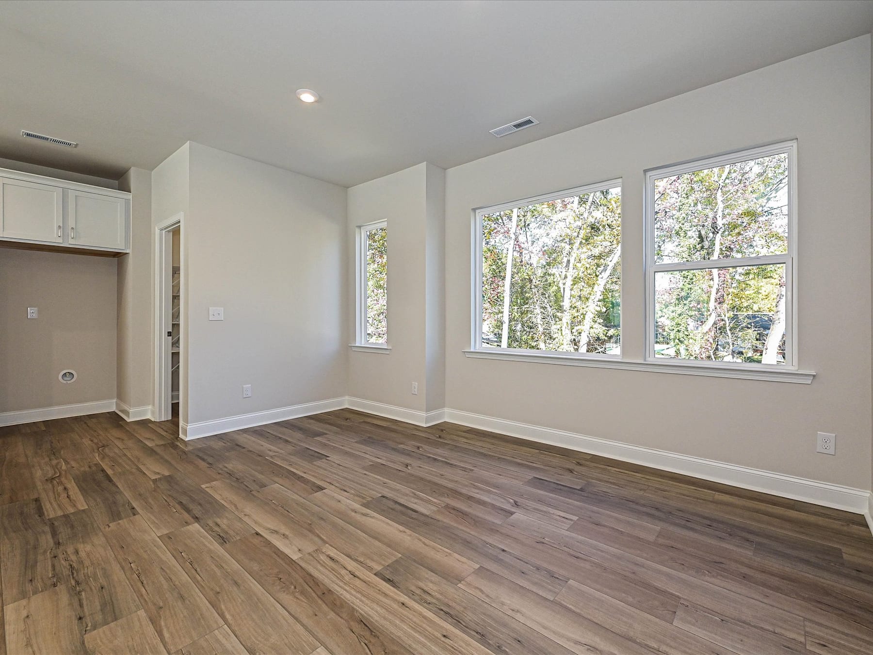 A spacious and well-lit room with hardwood floors, white walls, and large windows overlooking a wooded area.