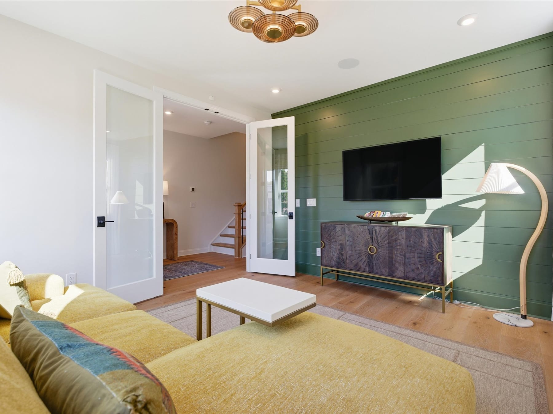A cozy and modern living room with a green accent wall, a TV stand, and a gold chandelier overhead, complemented by a plush yellow rug and a white coffee table in the foreground.