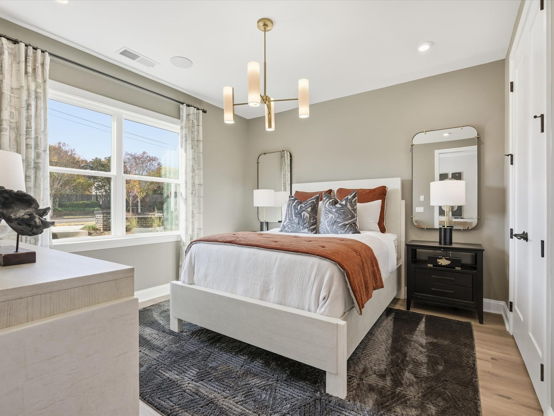 A cozy and well-appointed bedroom with a large window, a wooden bed frame, and a modern chandelier overhead, set against a neutral color palette and accented with textured bedding and decor.