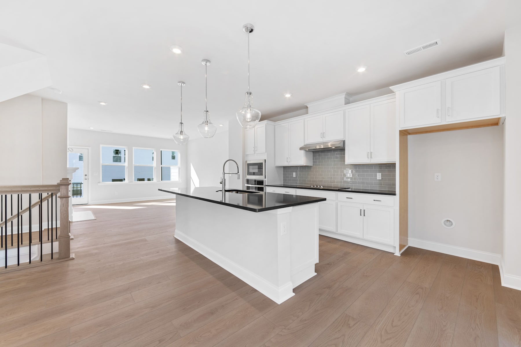 A modern, open-concept kitchen with white cabinets, a black countertop, and hardwood floors, leading into a spacious living area with high ceilings and pendant lighting.