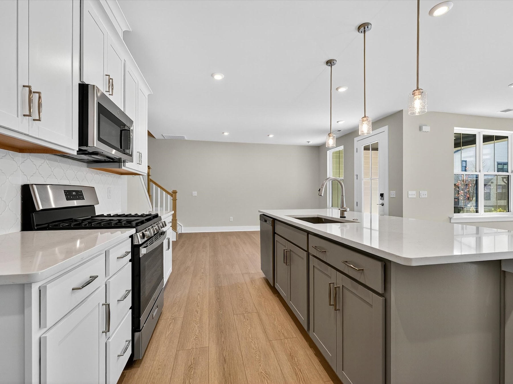 A modern, well-lit kitchen with white cabinets, stainless steel appliances, and a wooden floor, featuring a central island with pendant lighting.