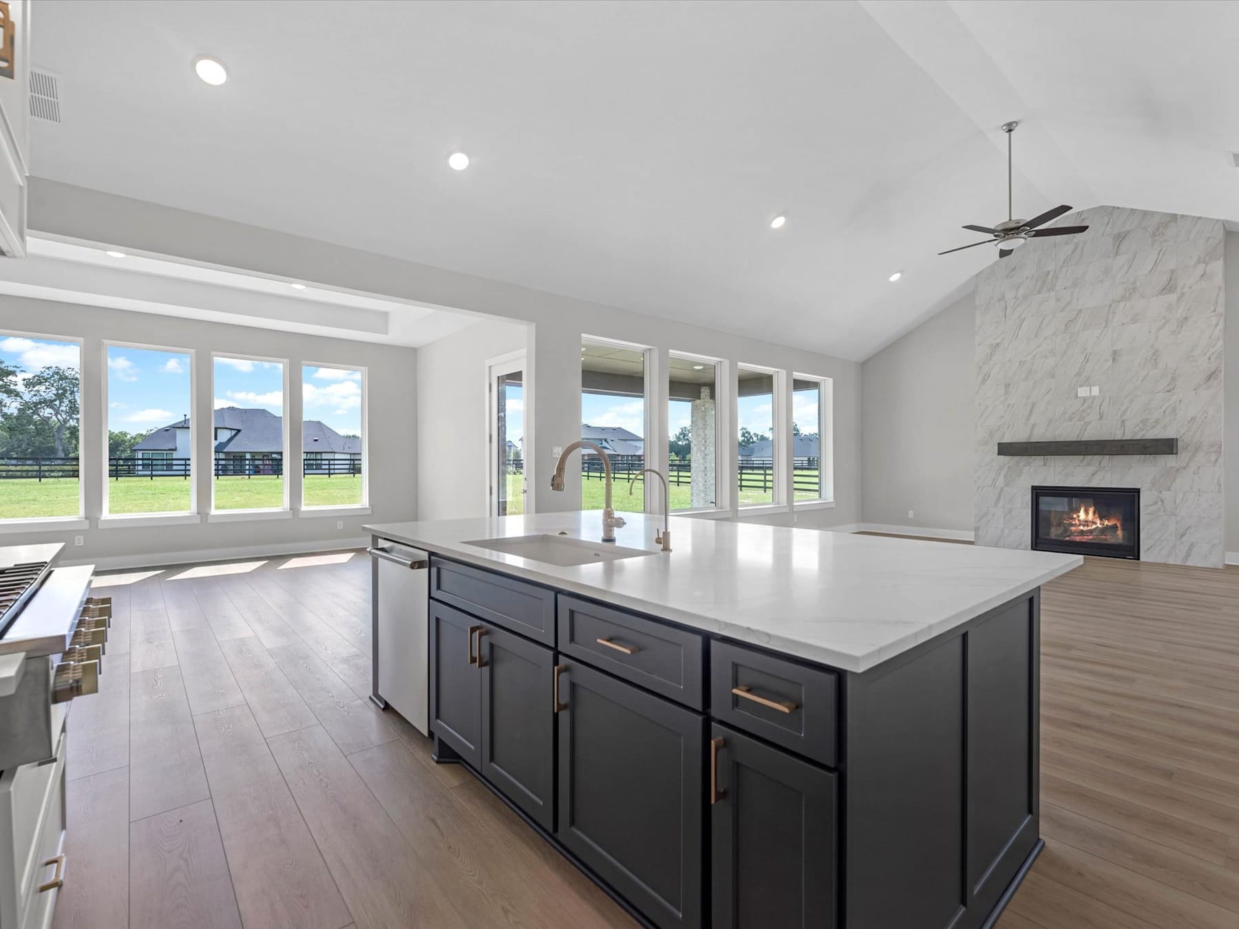 A spacious and modern kitchen with a large island, dark cabinets, and a fireplace in the background, surrounded by large windows offering a scenic view of the outdoor landscape.