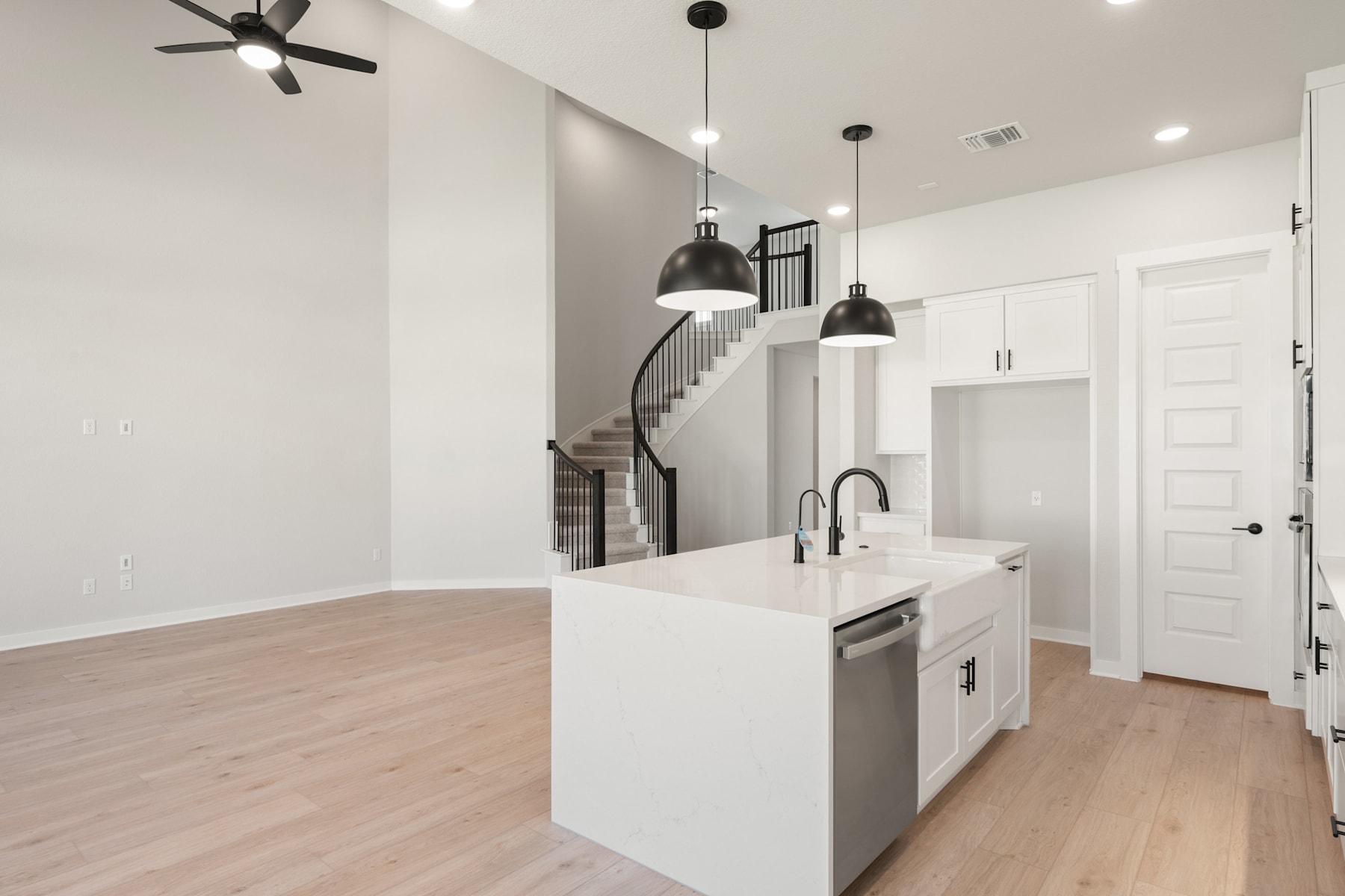 A modern, minimalist kitchen with white cabinets, a central island, and pendant lighting fixtures, set against a backdrop of a staircase and a white interior.