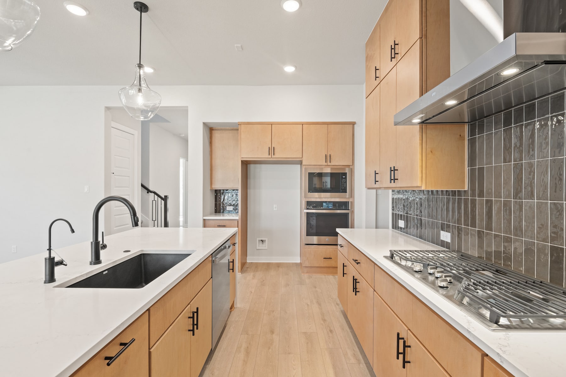 A modern, well-lit kitchen with light-colored wood cabinets, a white countertop, and stainless steel appliances, including a gas stove and a sink.
