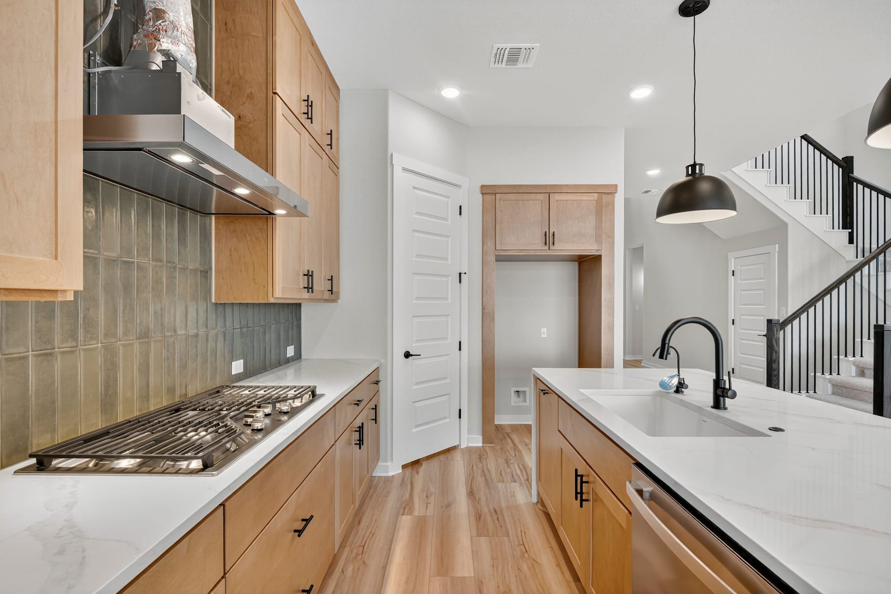 A modern and well-designed kitchen with light wood cabinets, a white countertop, and a tiled backsplash, leading into a hallway with a staircase visible in the background.