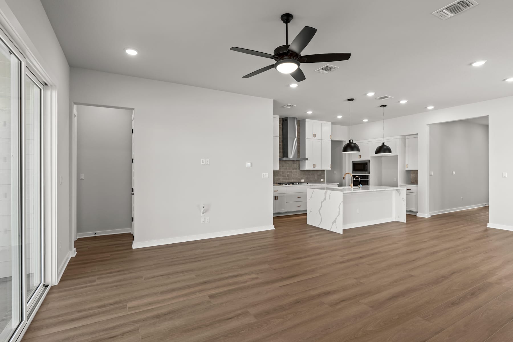 A spacious, open-concept living area with a kitchen visible in the background, featuring a ceiling fan, hardwood flooring, and a sliding glass door leading to an outdoor space.