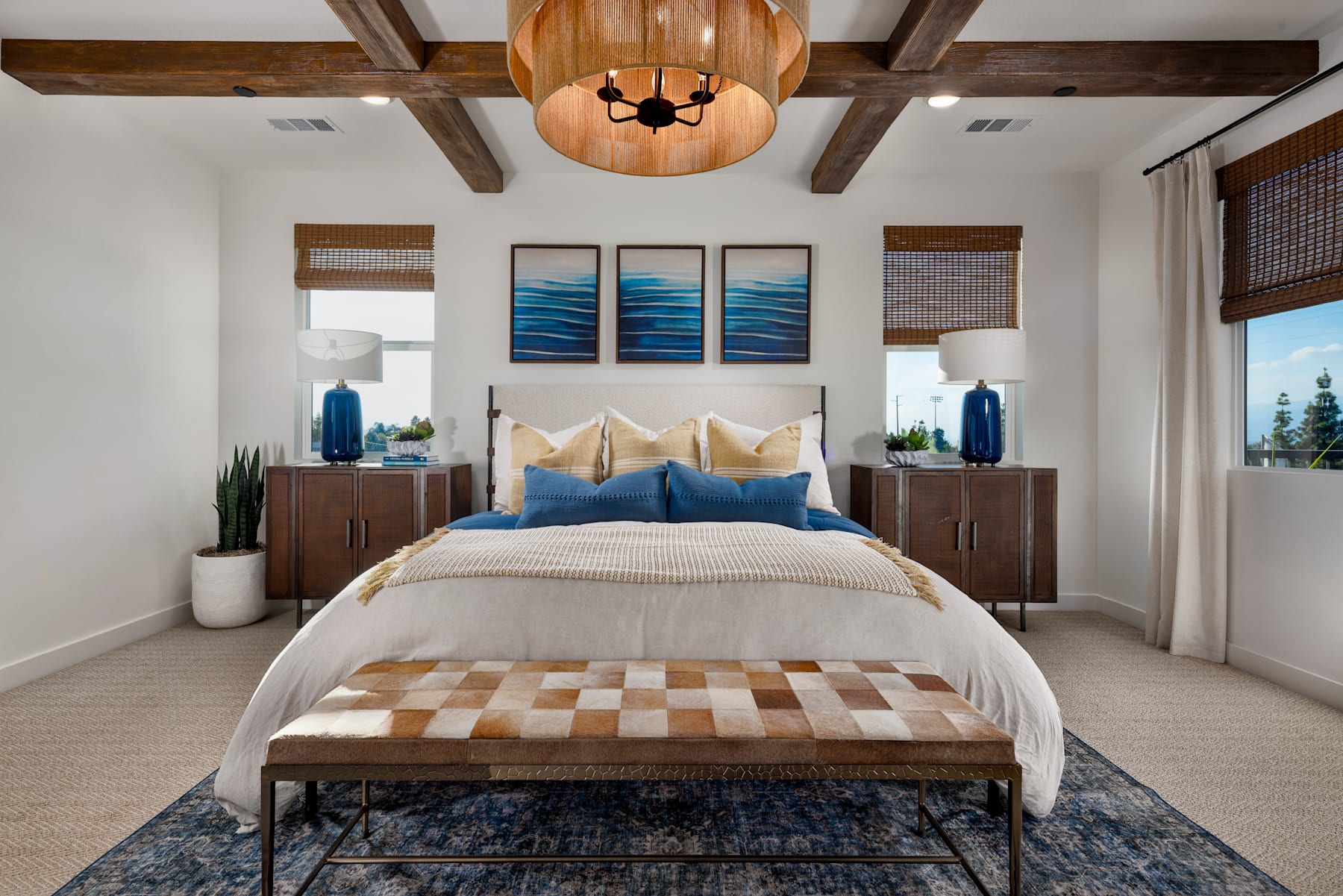 A cozy and inviting bedroom with wooden beams, a large pendant light, and artwork depicting ocean waves, complemented by a patterned bench and neutral bedding.