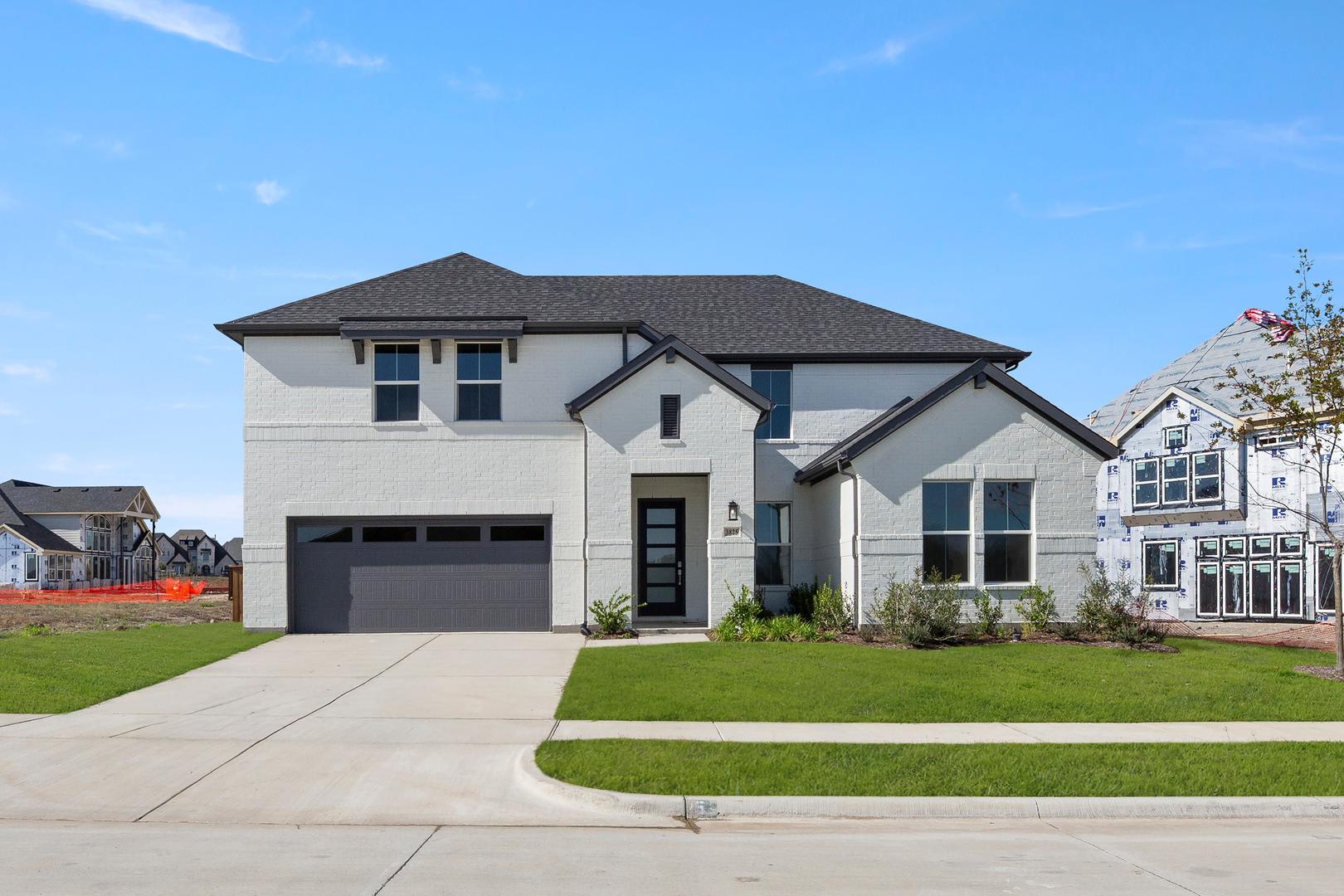 A large, modern two-story house with a gray roof and white exterior stands on a grassy lawn, surrounded by other houses in a residential neighborhood.