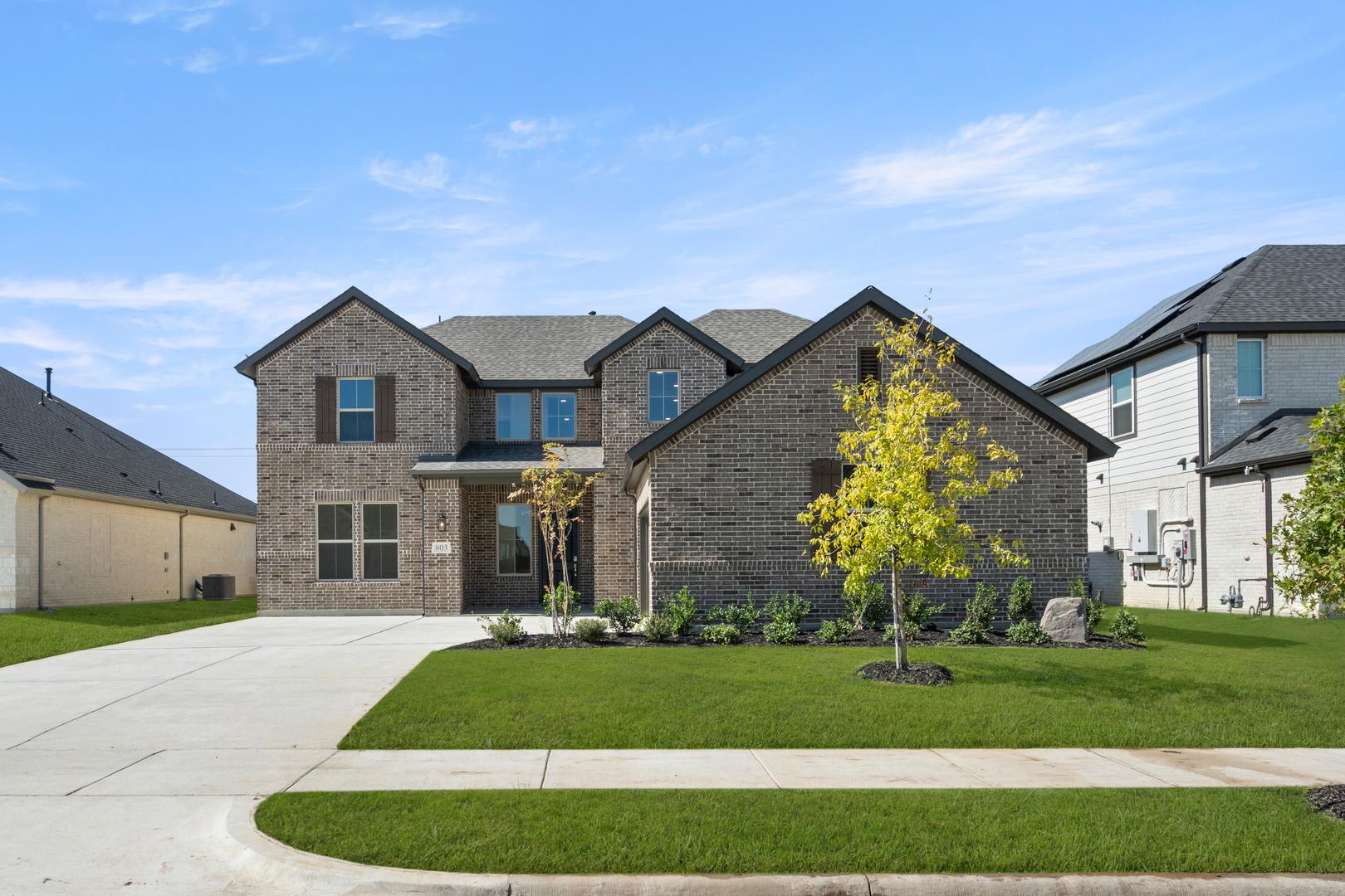 A two-story brick house with a well-manicured lawn, surrounded by other houses in a residential neighborhood, set against a clear blue sky with scattered clouds.