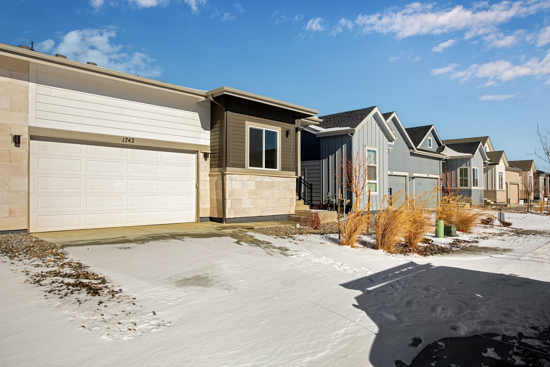 A row of modern, two-story townhouses with attached garages, set against a backdrop of a clear blue sky with scattered clouds, with a snowy path leading to the front doors.