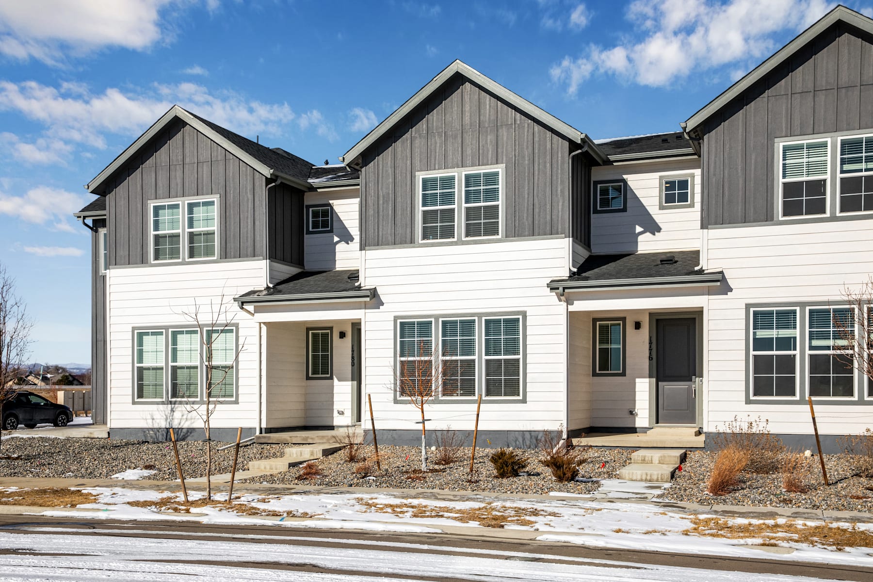 The image depicts a row of modern townhouses with a mix of gray and white siding, featuring gabled roofs and large windows. The foreground shows a snowy landscape with some landscaping elements, while the background features a clear blue sky with scattered clouds.