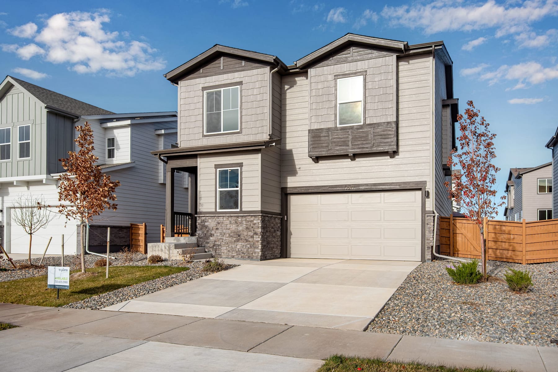 A two-story residential house with a garage, surrounded by a gravel driveway and landscaping, set against a blue sky with scattered clouds.