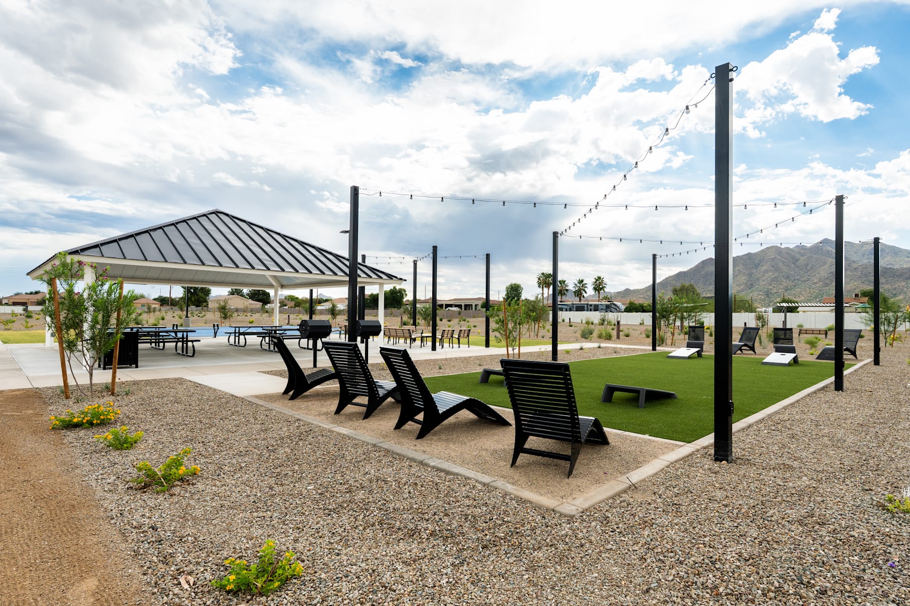 A covered outdoor seating area with black chairs and tables, surrounded by a gravel surface and lush greenery, set against a backdrop of a cloudy sky.