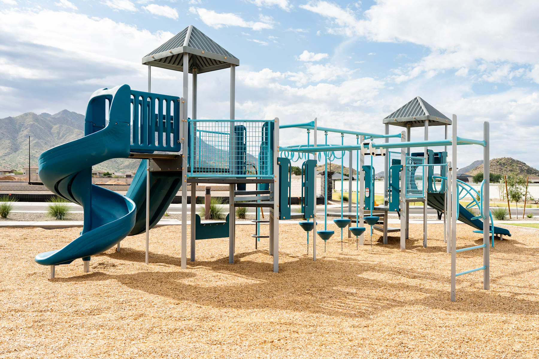 A colorful and modern playground structure with slides, ladders, and platforms stands in a sandy area, surrounded by a mountainous landscape under a cloudy sky.