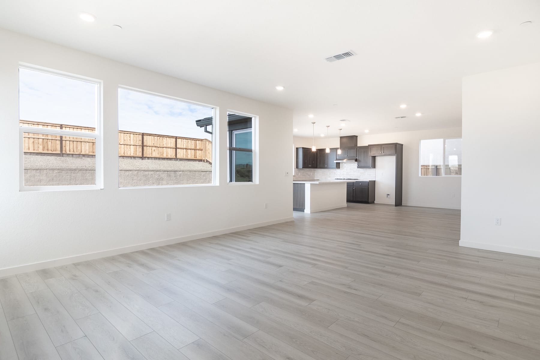 A spacious, modern living room with a sleek kitchen visible in the background, featuring large windows that allow natural light to flood the space.