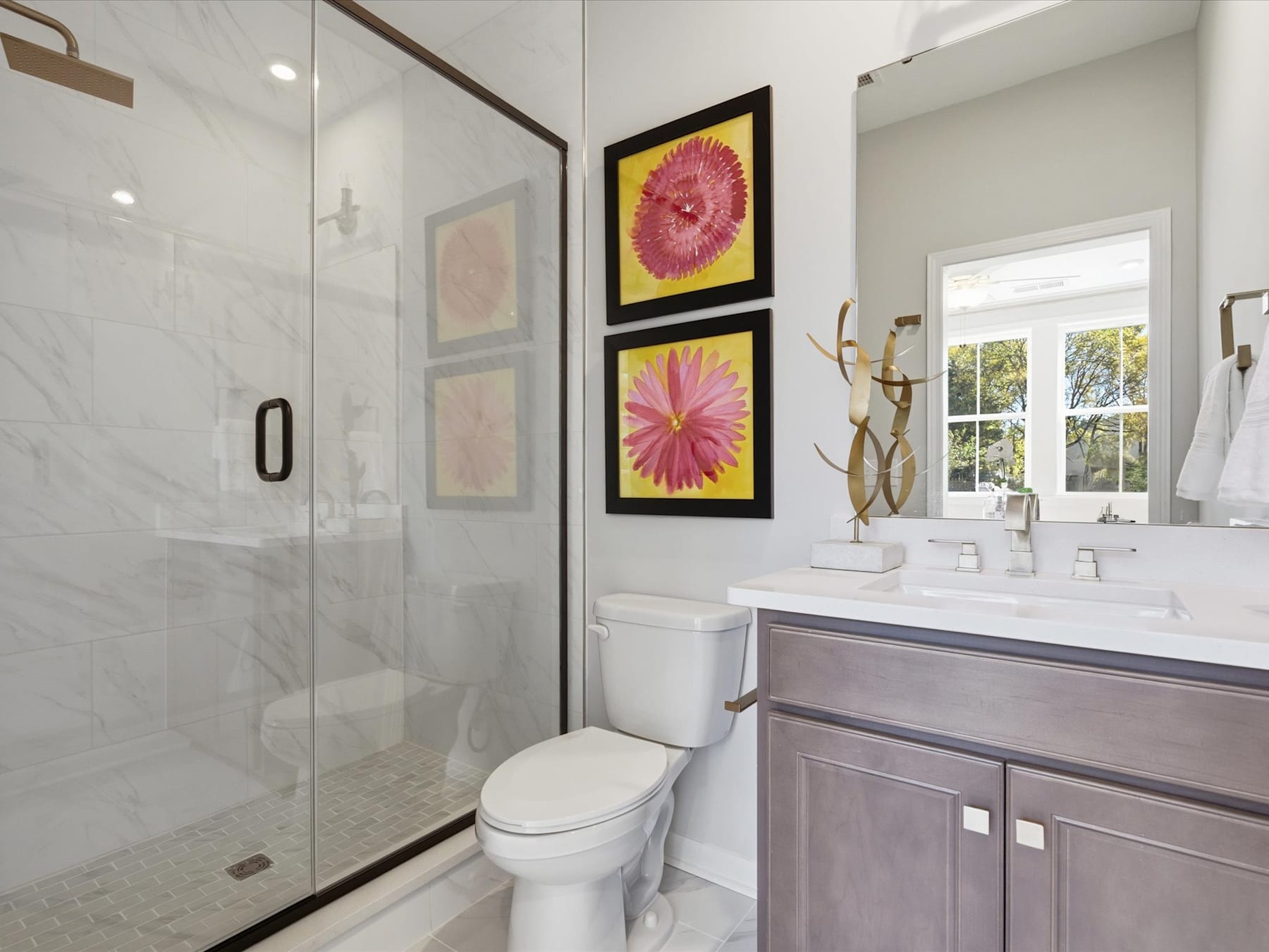A modern bathroom with a glass shower enclosure, a vanity with a white countertop, and framed artwork featuring vibrant pink flowers on the wall.