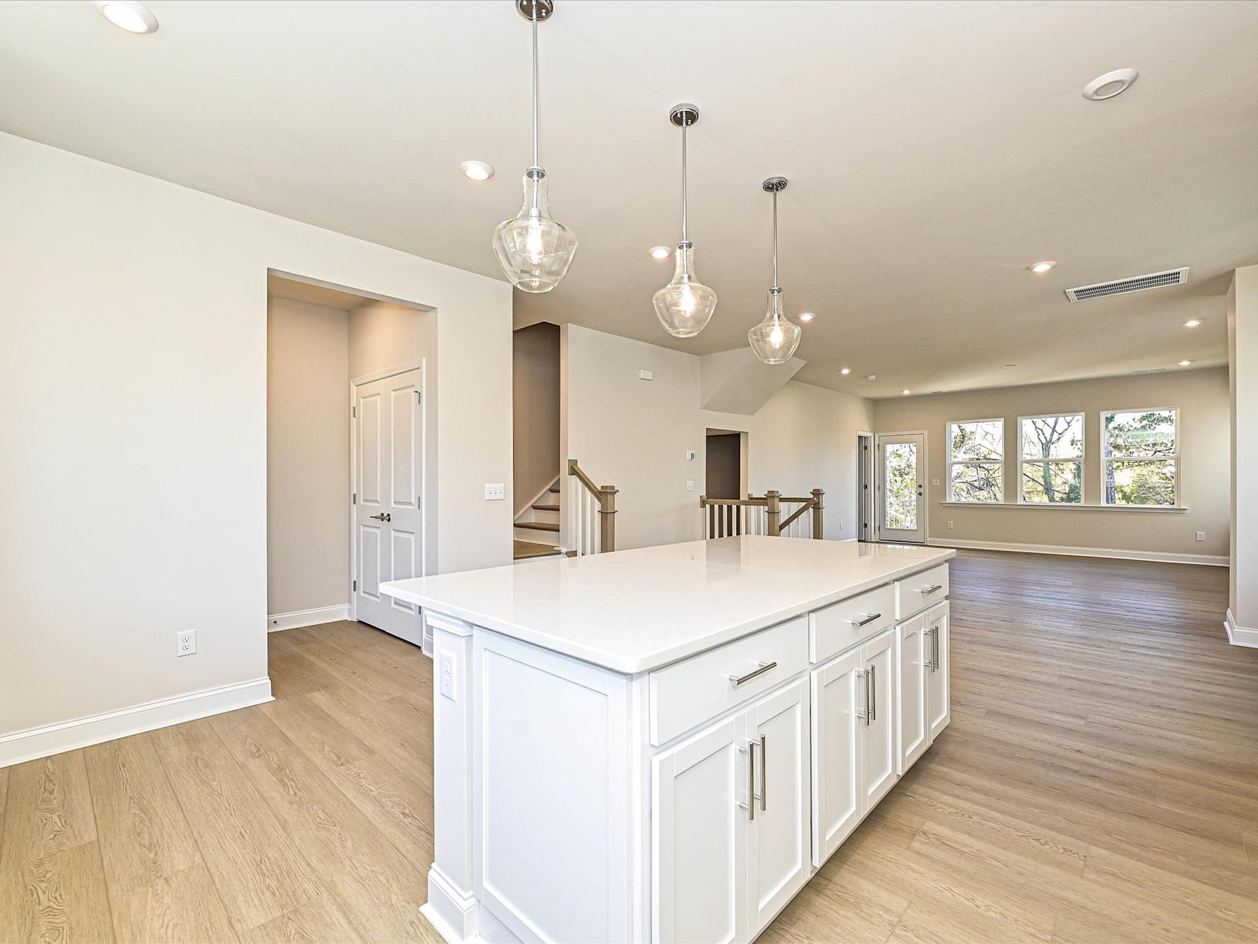 A spacious and modern kitchen with a large white island, pendant lighting, and hardwood floors, set against a backdrop of an open living area with windows.