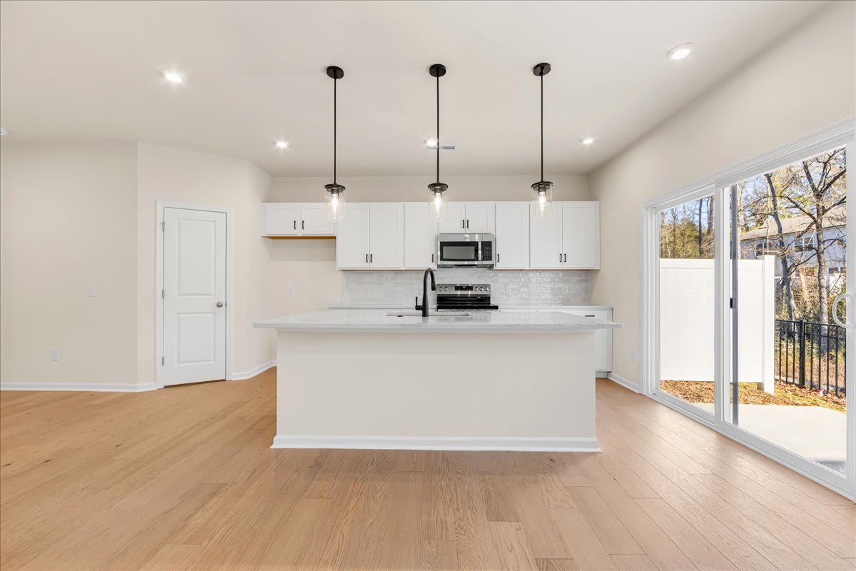 A modern, open-concept kitchen with white cabinets, a central island, and hardwood floors, surrounded by large windows that allow natural light to flood the space.