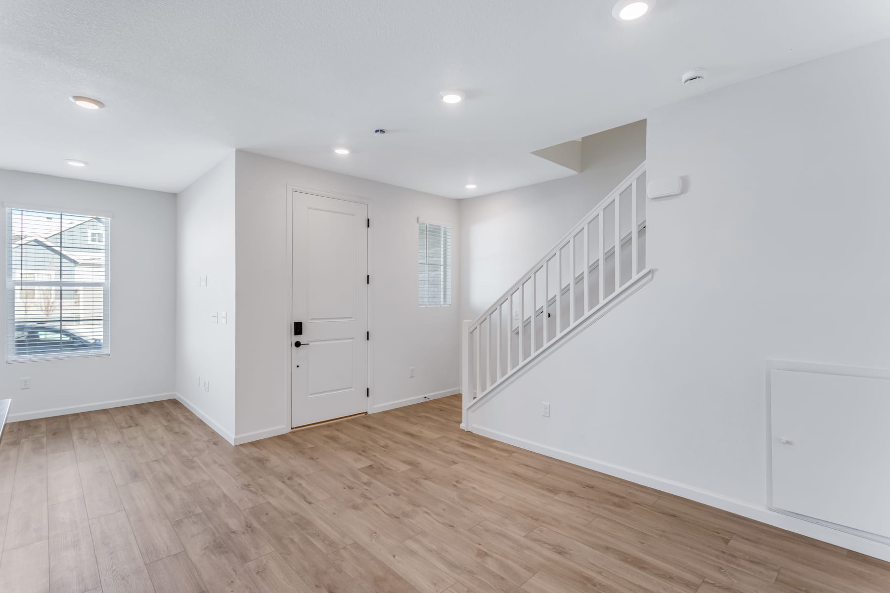 A bright and spacious interior with white walls, a wooden floor, and a staircase leading to an upper level. The room appears to be a hallway or entryway, with a door visible in the background.