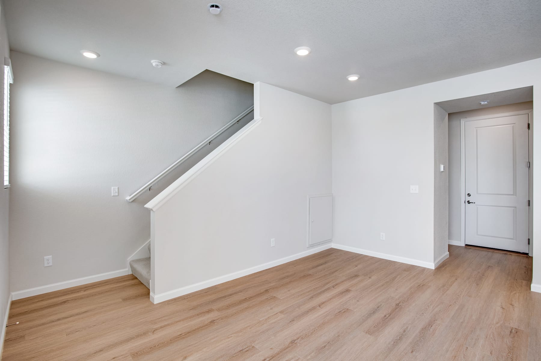 A spacious, well-lit room with white walls, a wooden floor, and a staircase leading to an upper level. The room appears to be a living or dining area, with a door visible in the background.