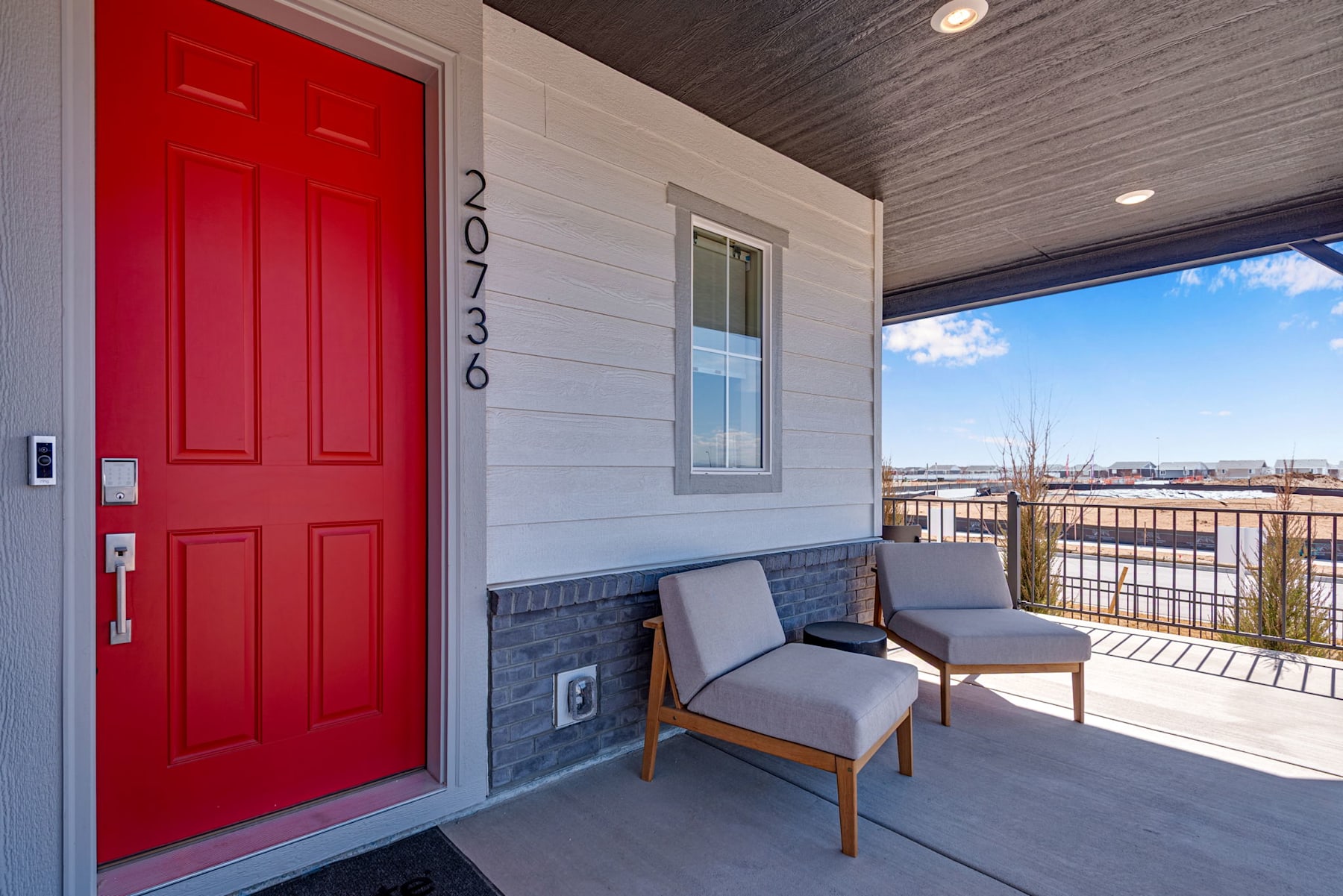 A bright red front door stands out against the gray siding of the house, with a small porch featuring a couple of chairs overlooking a scenic outdoor landscape in the background.