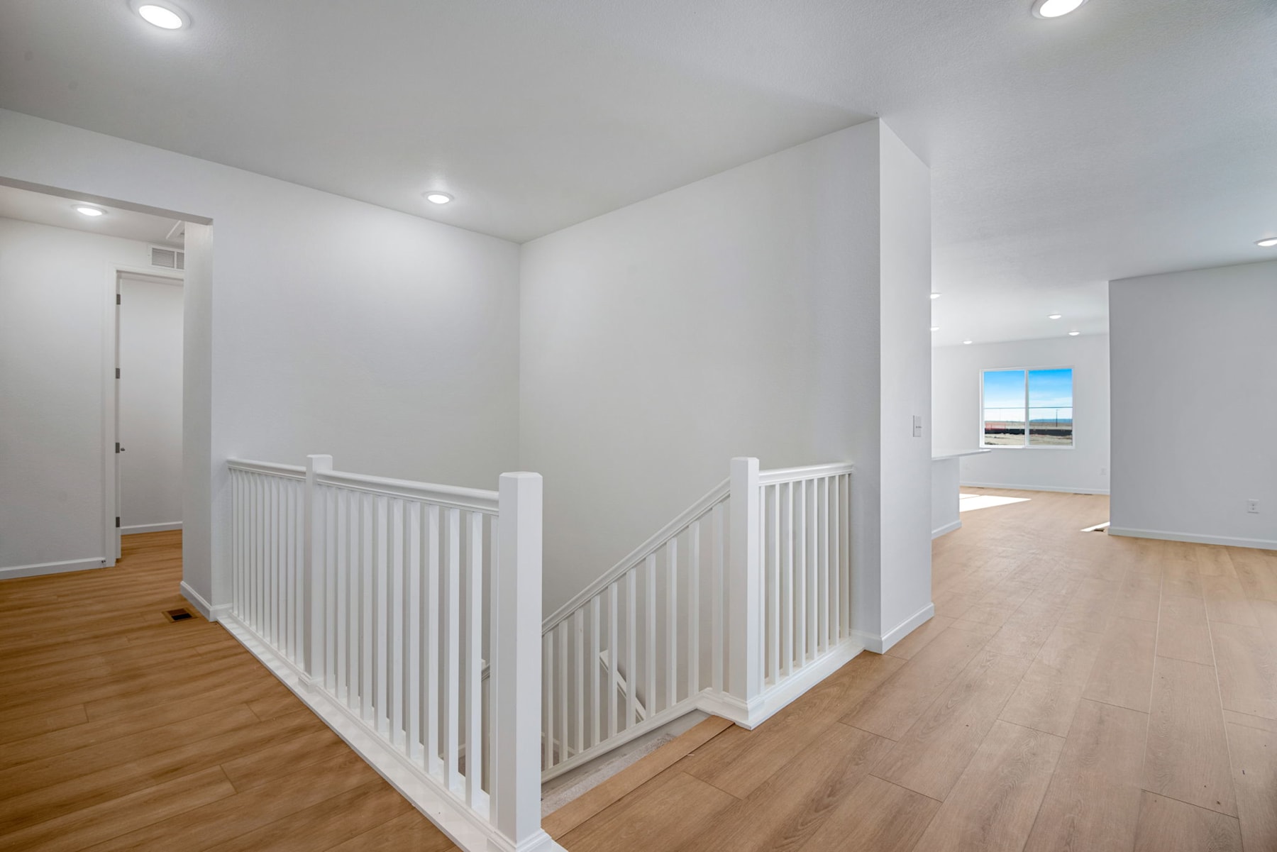 A bright, open-concept hallway with white walls, wooden floors, and a staircase with white railings leading to another room.