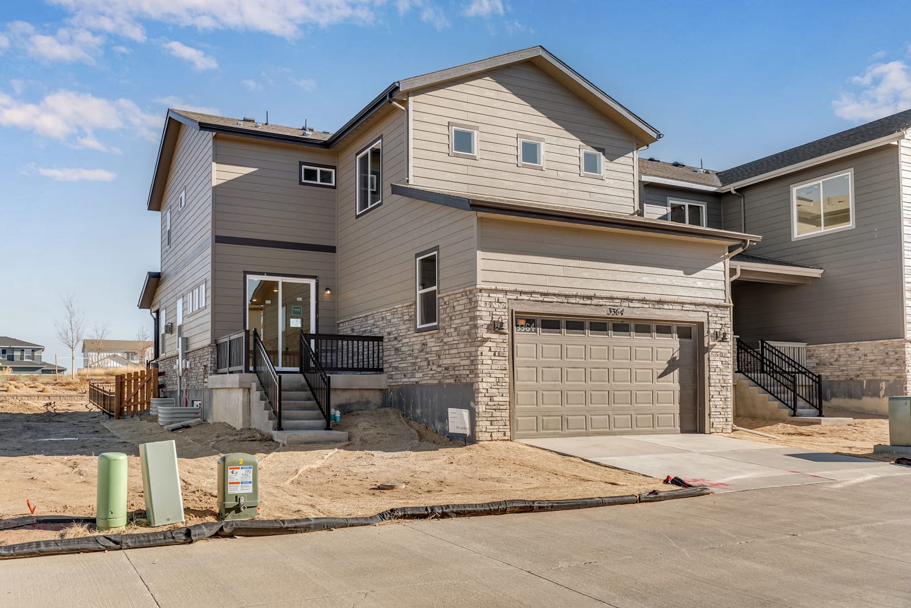 A newly constructed two-story residential house with a garage, situated on a sandy lot with construction materials in the foreground, against a backdrop of a clear blue sky with scattered clouds.
