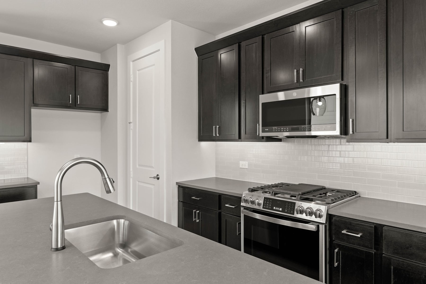 A modern kitchen with dark cabinets, a stainless steel sink, and a gas stove in the foreground, set against a white tiled backsplash and walls.
