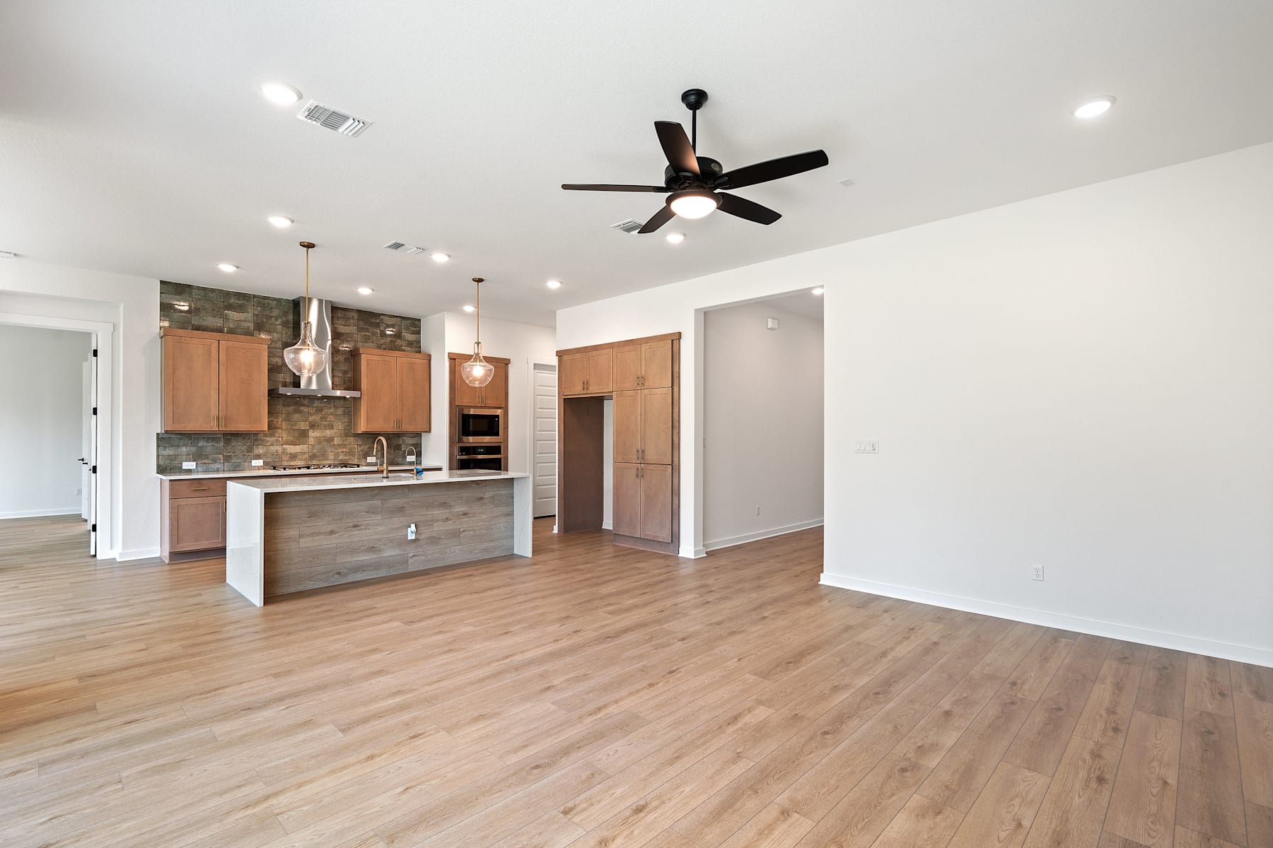 A modern, open-concept kitchen and living space with hardwood floors, a ceiling fan, and a mix of light and dark wood cabinetry.