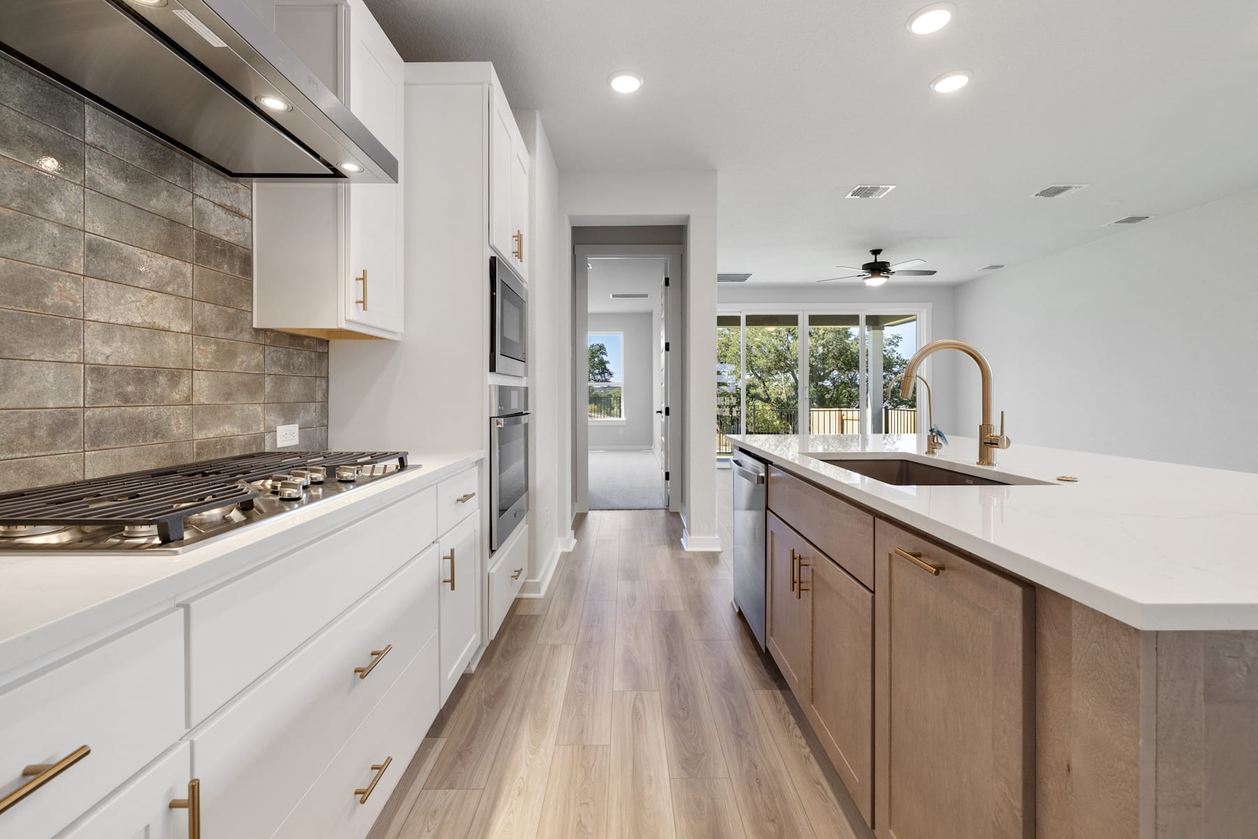 A modern, well-lit kitchen with white cabinets, a tile backsplash, and a wooden countertop, leading to a hallway with a view of the outdoors.