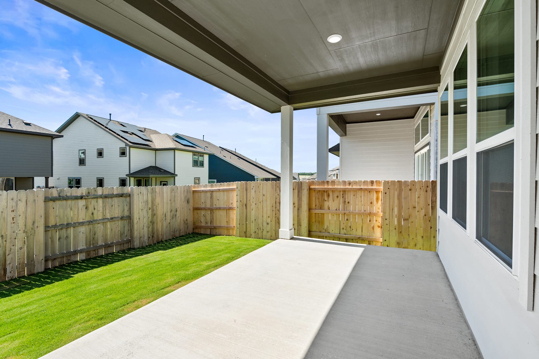 A covered patio with a concrete walkway leading to a grassy backyard, surrounded by a wooden fence and neighboring houses in the background.