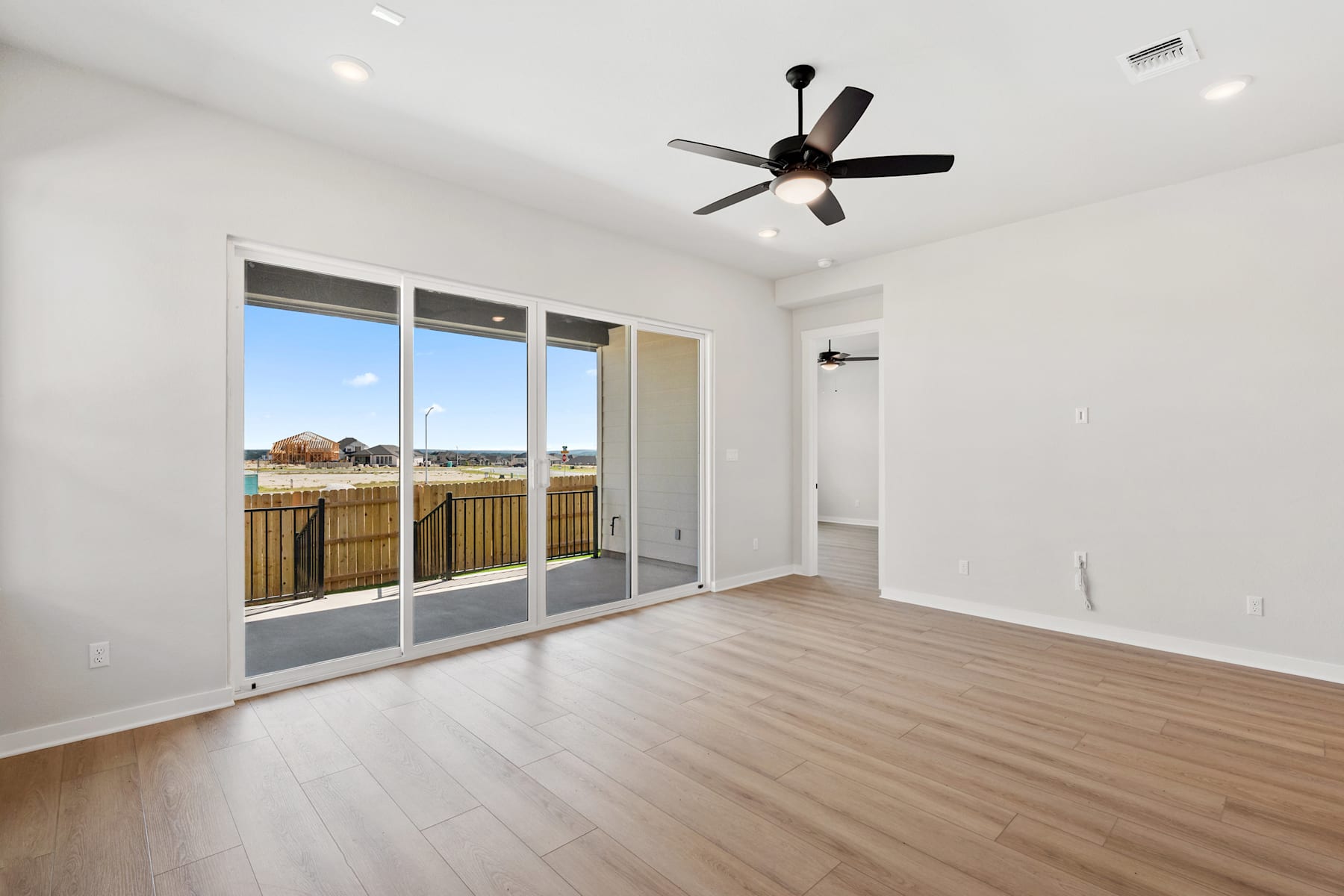 A spacious, well-lit room with a ceiling fan, sliding glass doors leading to a balcony, and a hardwood floor.