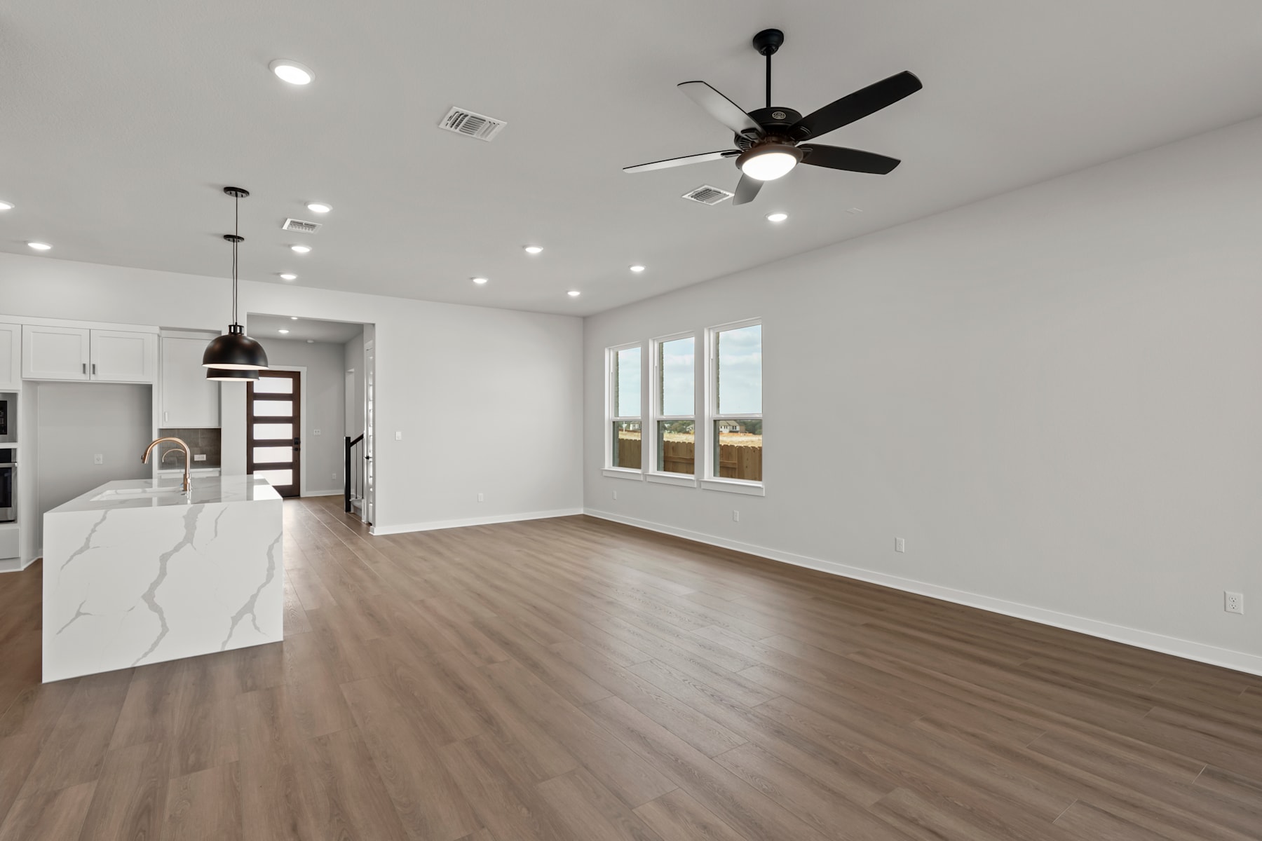 A spacious, well-lit room with a ceiling fan, hardwood floors, and a kitchen visible in the background.