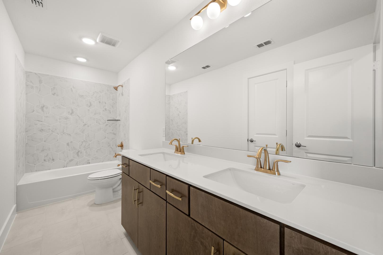 A modern and spacious bathroom with a double vanity, white marble countertops, and gold-toned fixtures, set against a backdrop of white walls and recessed lighting.