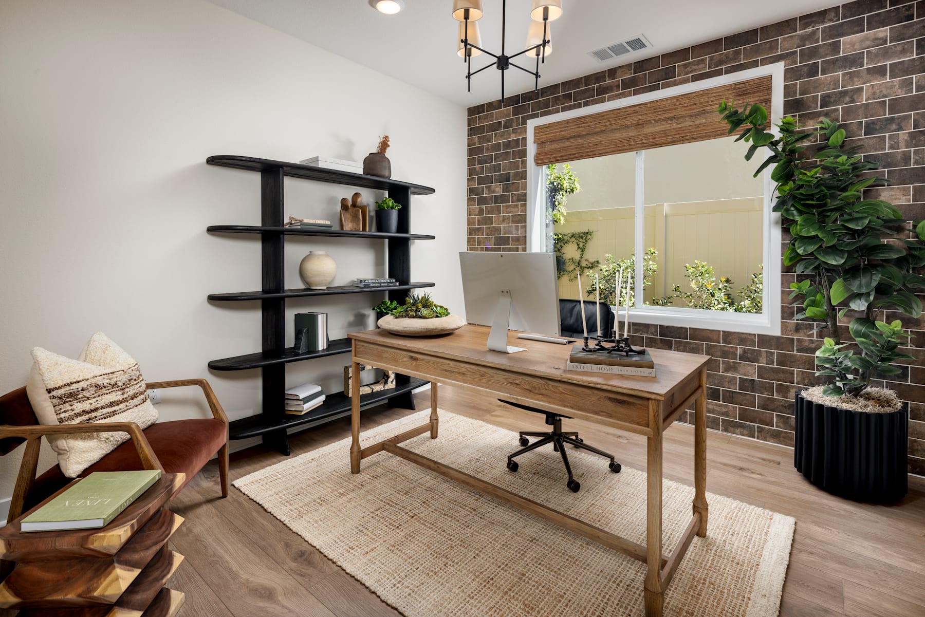 A cozy and well-designed home office space with a wooden desk, shelving unit, and various decorative elements, set against a brick wall backdrop and complemented by natural lighting and potted plants.