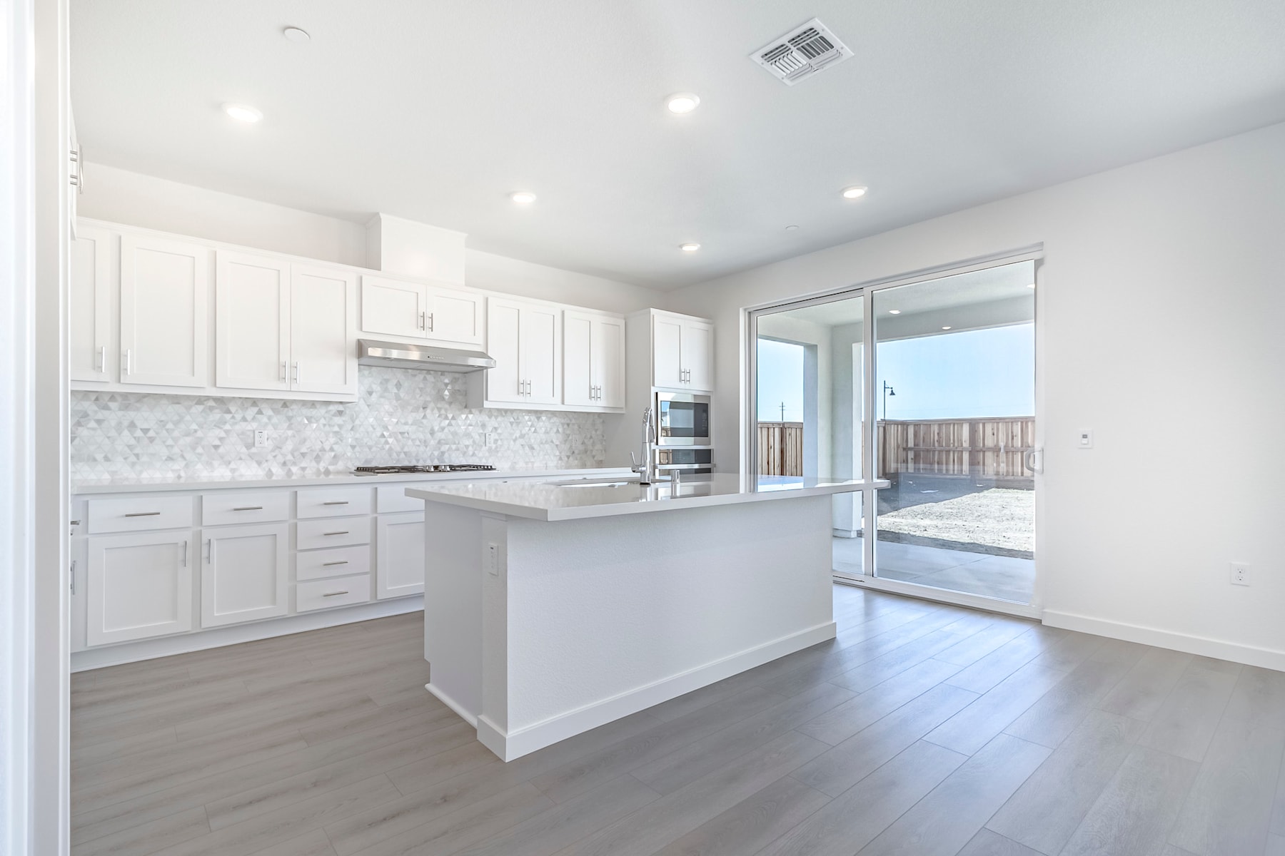 A modern, bright kitchen with white cabinets, a marble backsplash, and hardwood floors, leading to a sliding glass door that opens to an outdoor area.