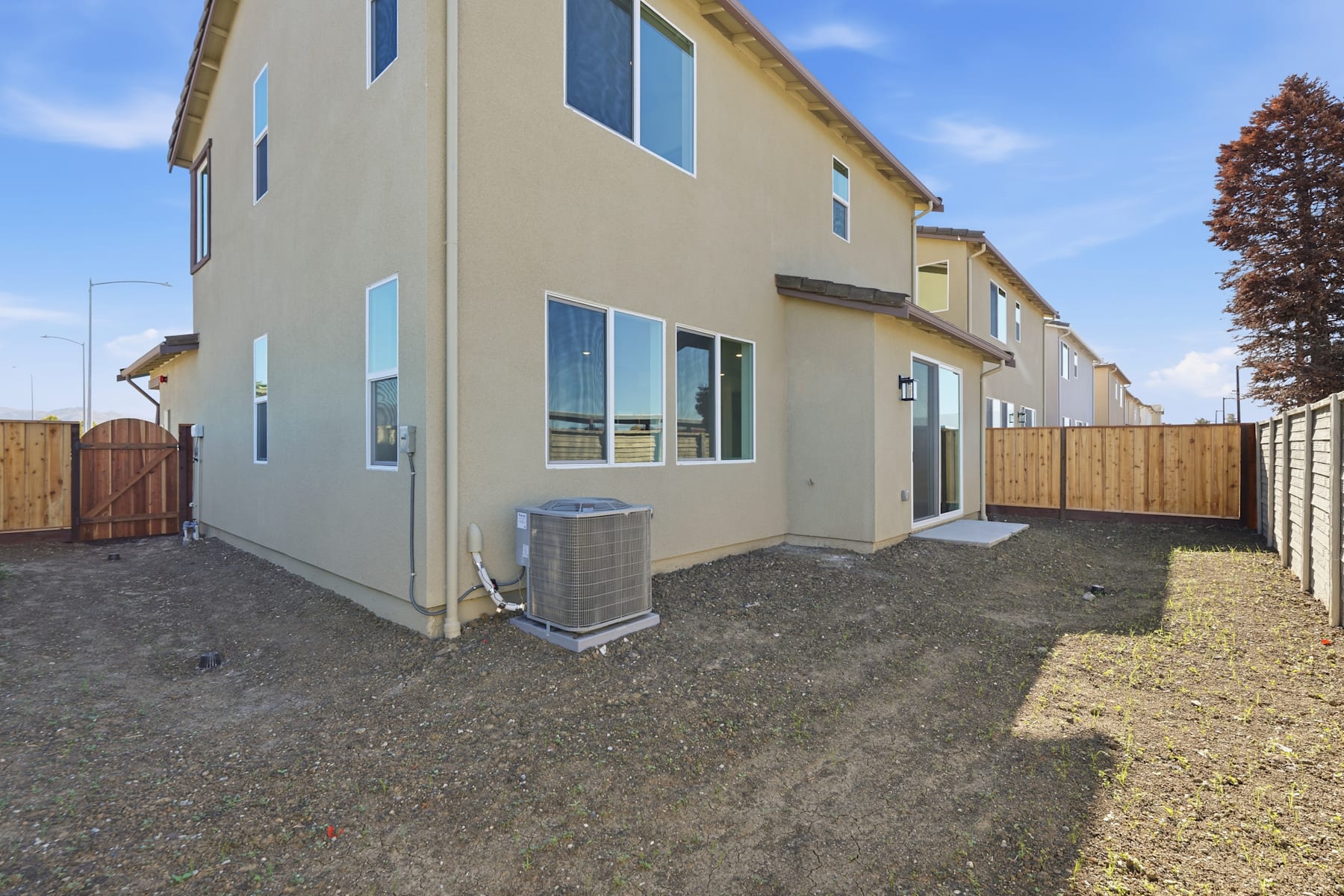 The image shows a row of modern, two-story townhouses with beige exterior walls and large windows. In the foreground, there is a grassy area with a concrete path leading to the houses, and a wooden fence separates the properties.
