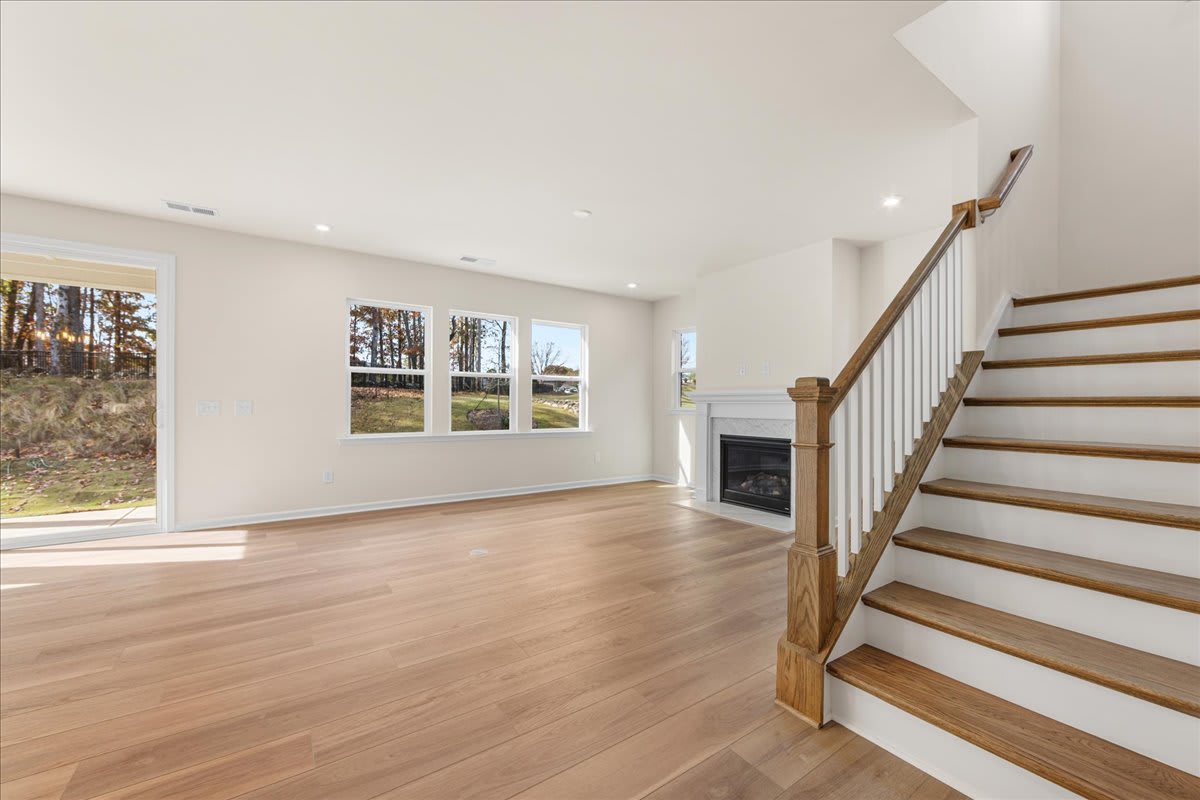 A spacious and bright living room with hardwood floors, a fireplace, and a staircase leading to the upper level. Large windows provide a view of the surrounding natural landscape.